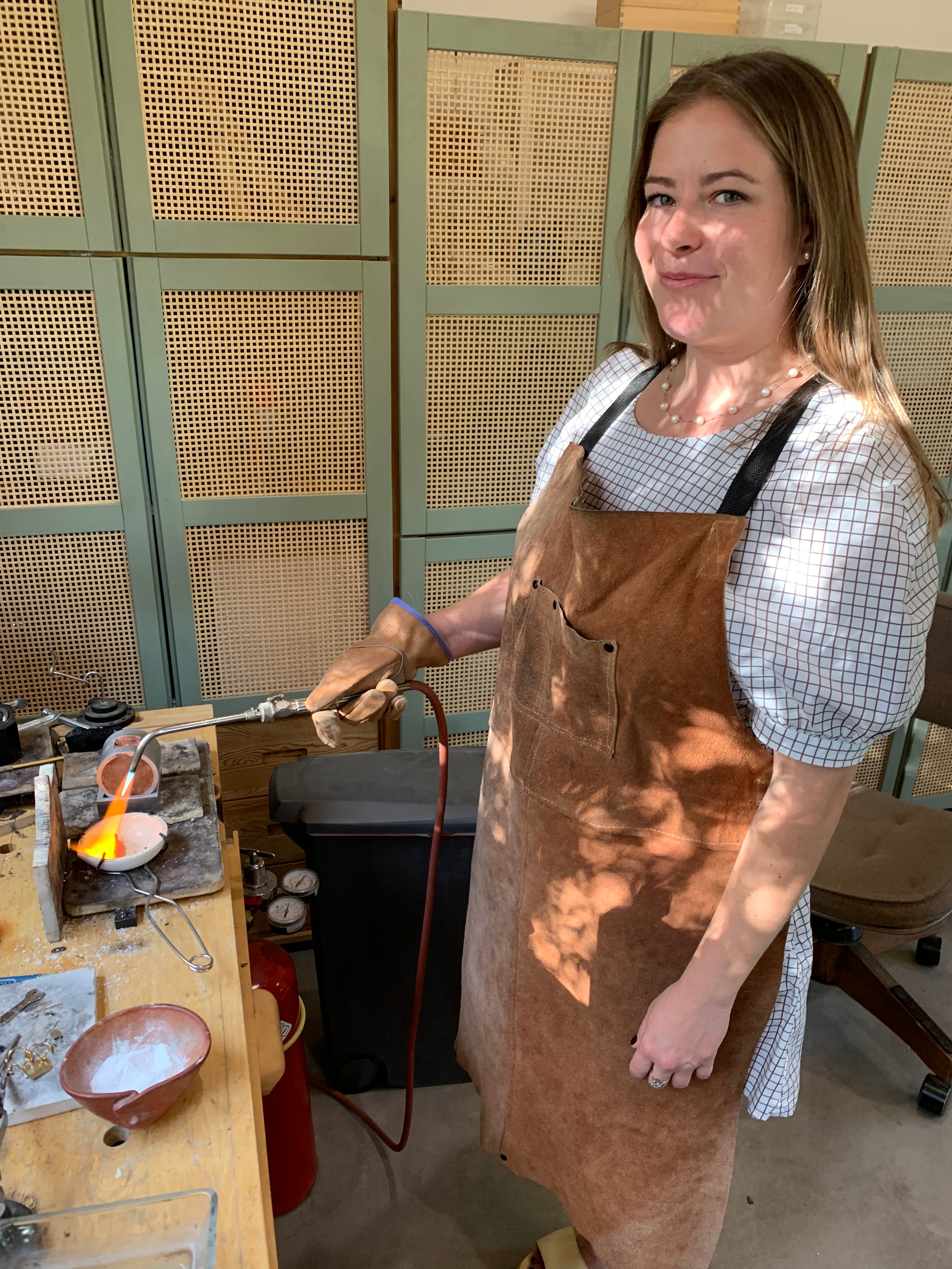 Person wearing a brown apron in a workshop setting with a jewelry making torch.